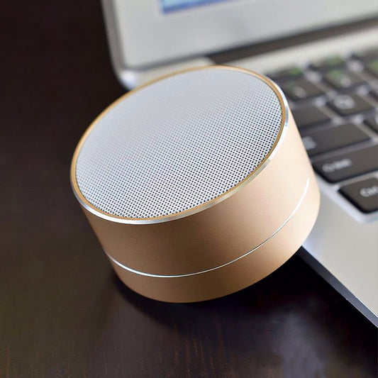 A gold-colored mini Bluetooth speaker placed on a wooden surface next to a laptop, indicating portability and use with electronic devices.