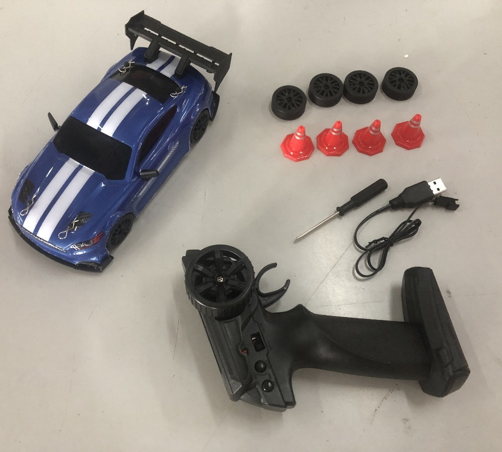 Close-up of blue RC drift car with controller, USB charger, screwdriver, spare tires, and traffic cones.