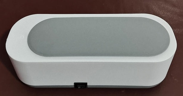 Modern white and gray ultrasonic cleaning device placed on a dark brown surface, featuring a central power switch.