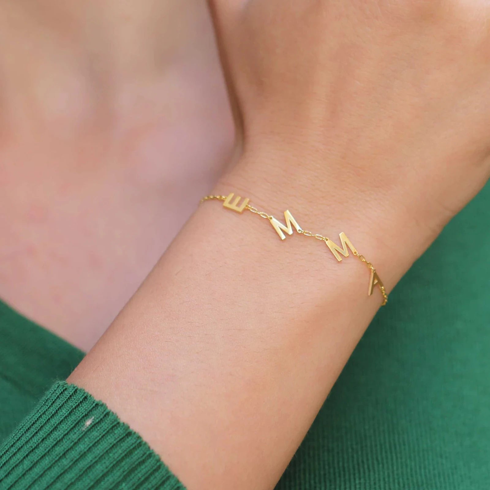 Gold bracelet with 'MAMA' letters on a person's wrist against a green background