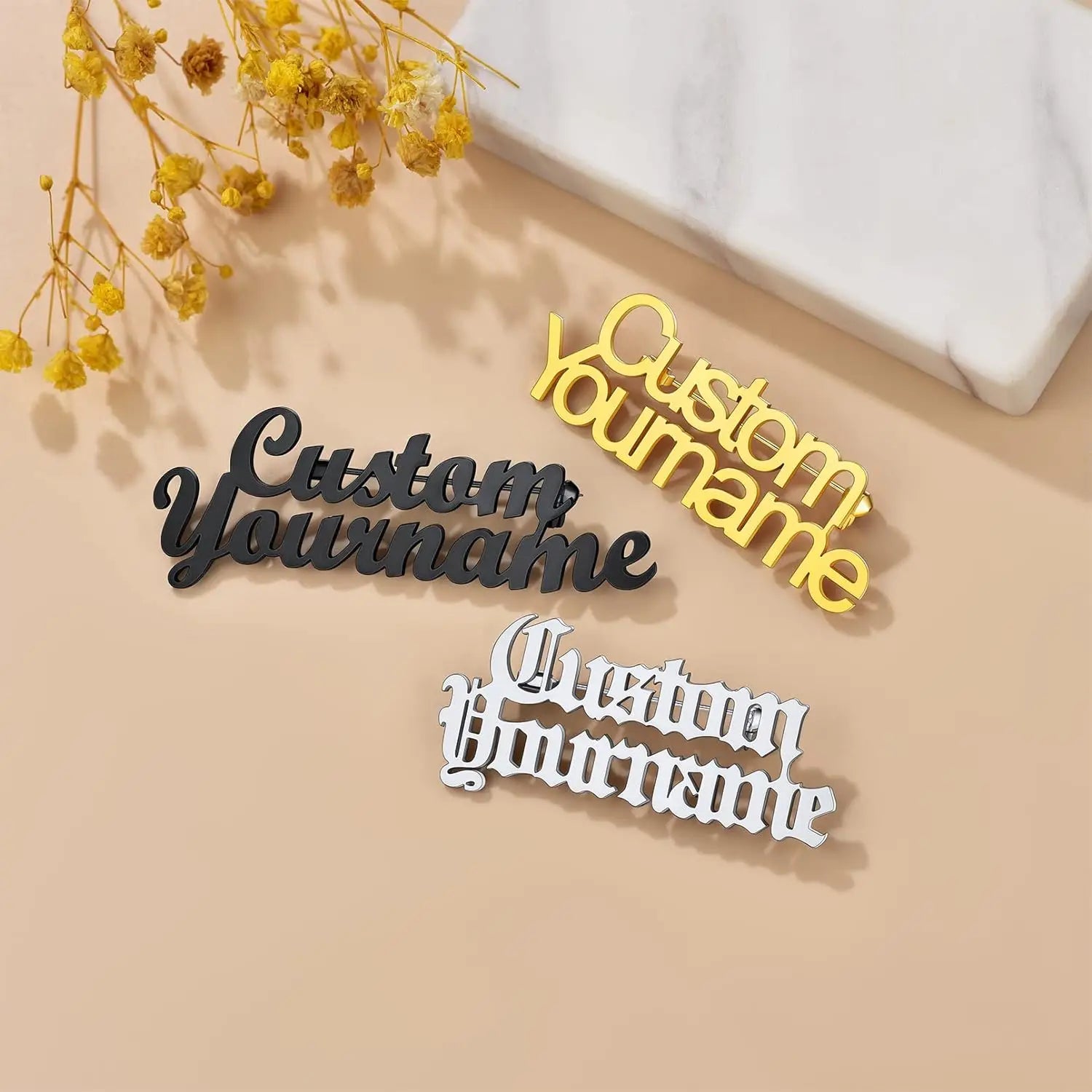 Three customizable name plates in black, yellow, and white on a beige surface with dried yellow flowers.