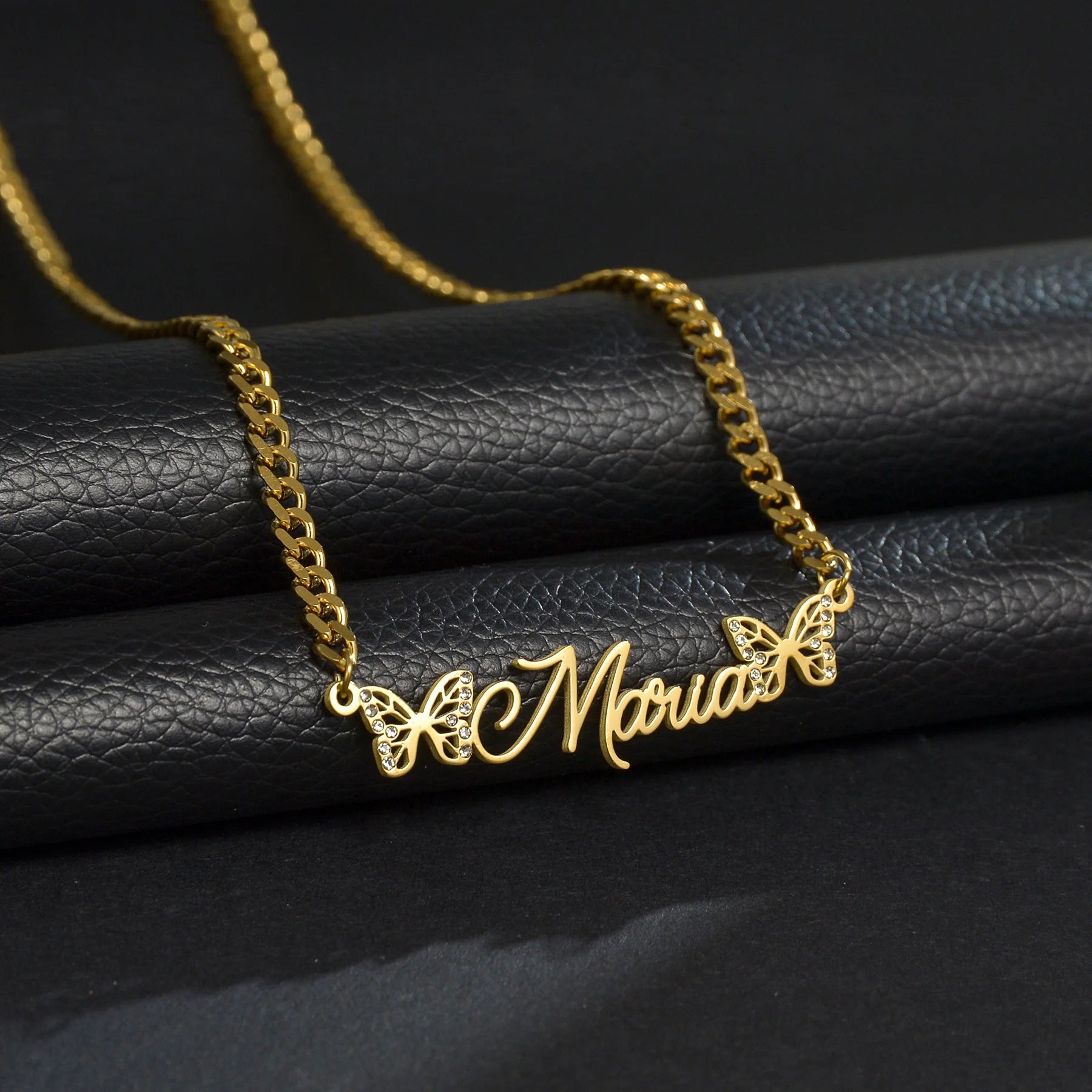 Gold necklace with 'Maria' and butterfly designs on a black leather surface