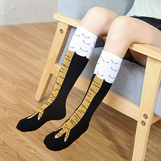 Person wearing black novelty knee-high socks with a fun eagle leg design, featuring yellow talons on the feet and white feather patterns at the top, seated on a wooden chair.