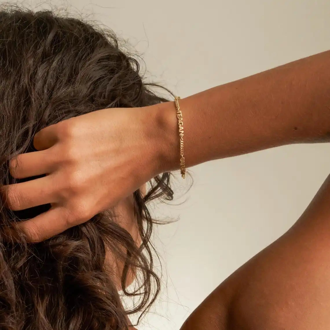 Person with a gold bracelet on their wrist, touching their hair against a neutral background
