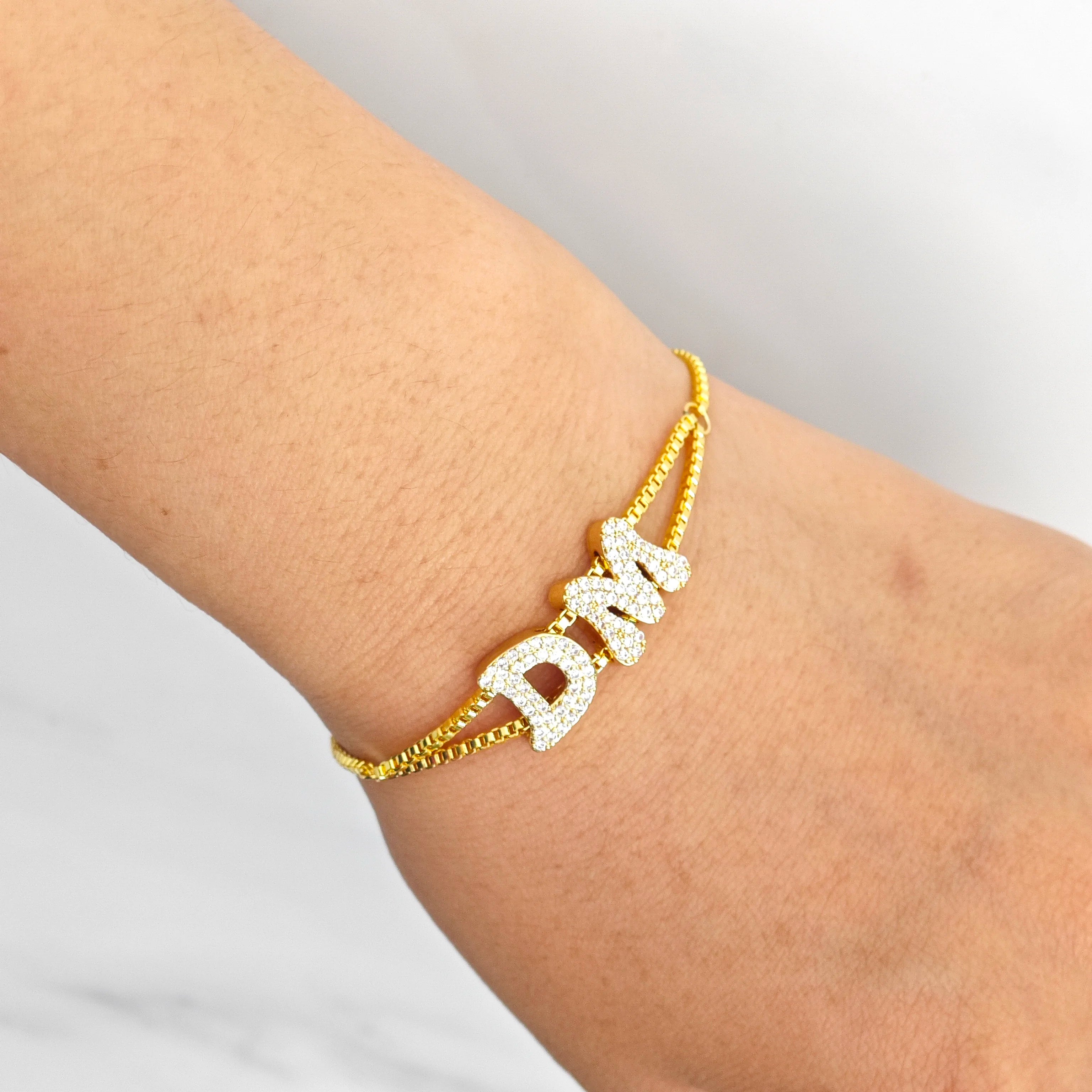 Gold bracelet with 'DAM' letters on a wrist against a light gray background