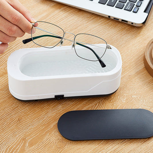 A pair of eyeglasses being cleaned by a white ultrasonic cleaner placed on a wooden table.