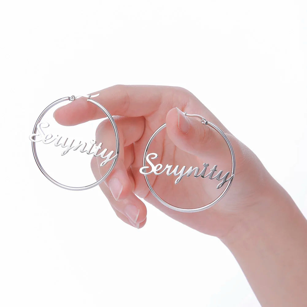 Silver hoop earrings with 'Serynity' engraving held by a hand on a white background