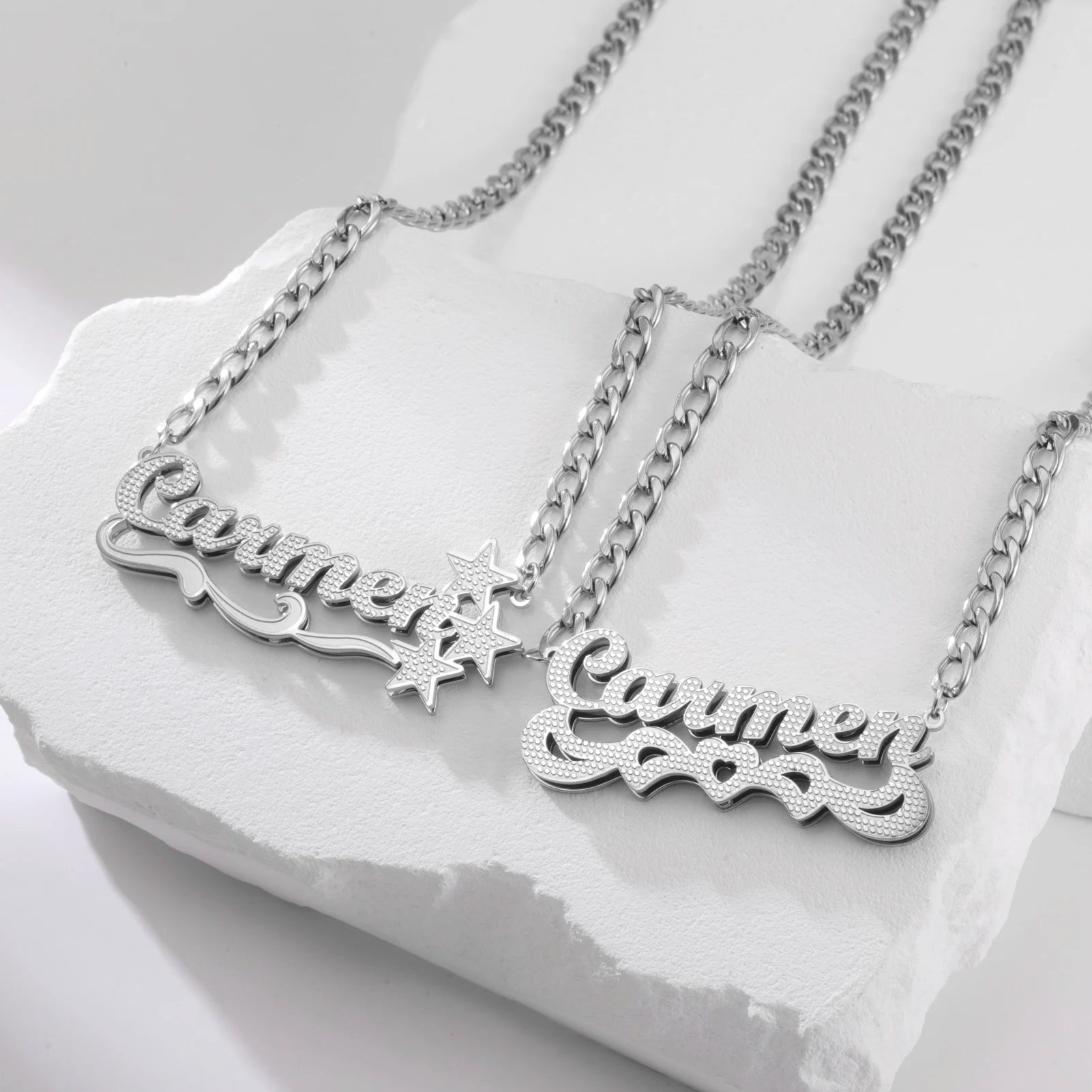 Two silver nameplate necklaces on a white stone surface