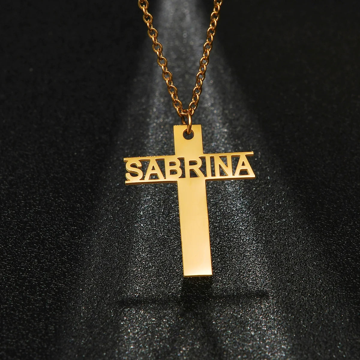 Gold cross pendant with 'Sabrina' engraving on a black background